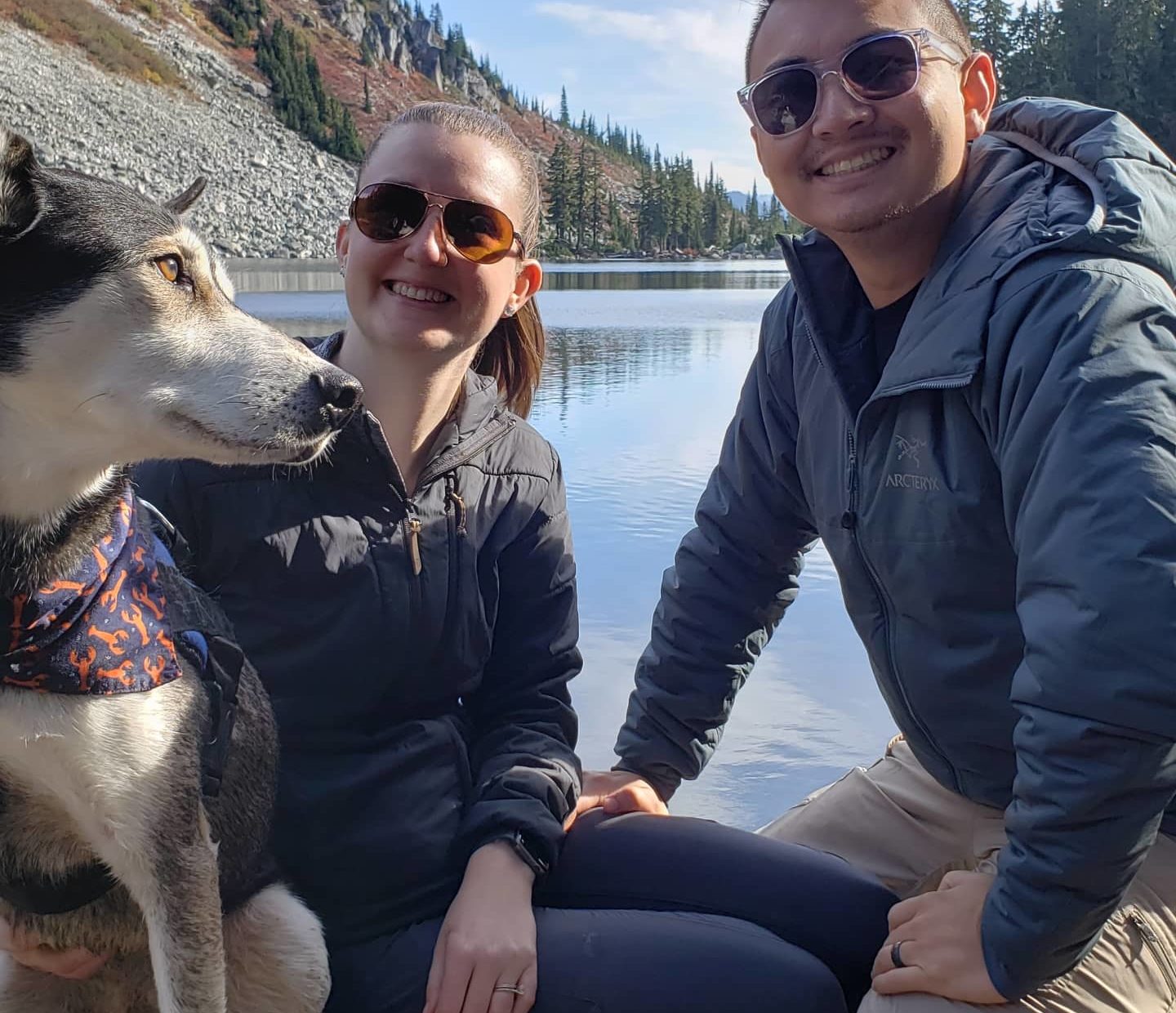 couple poses with their dog in front of lake and mountain landscape