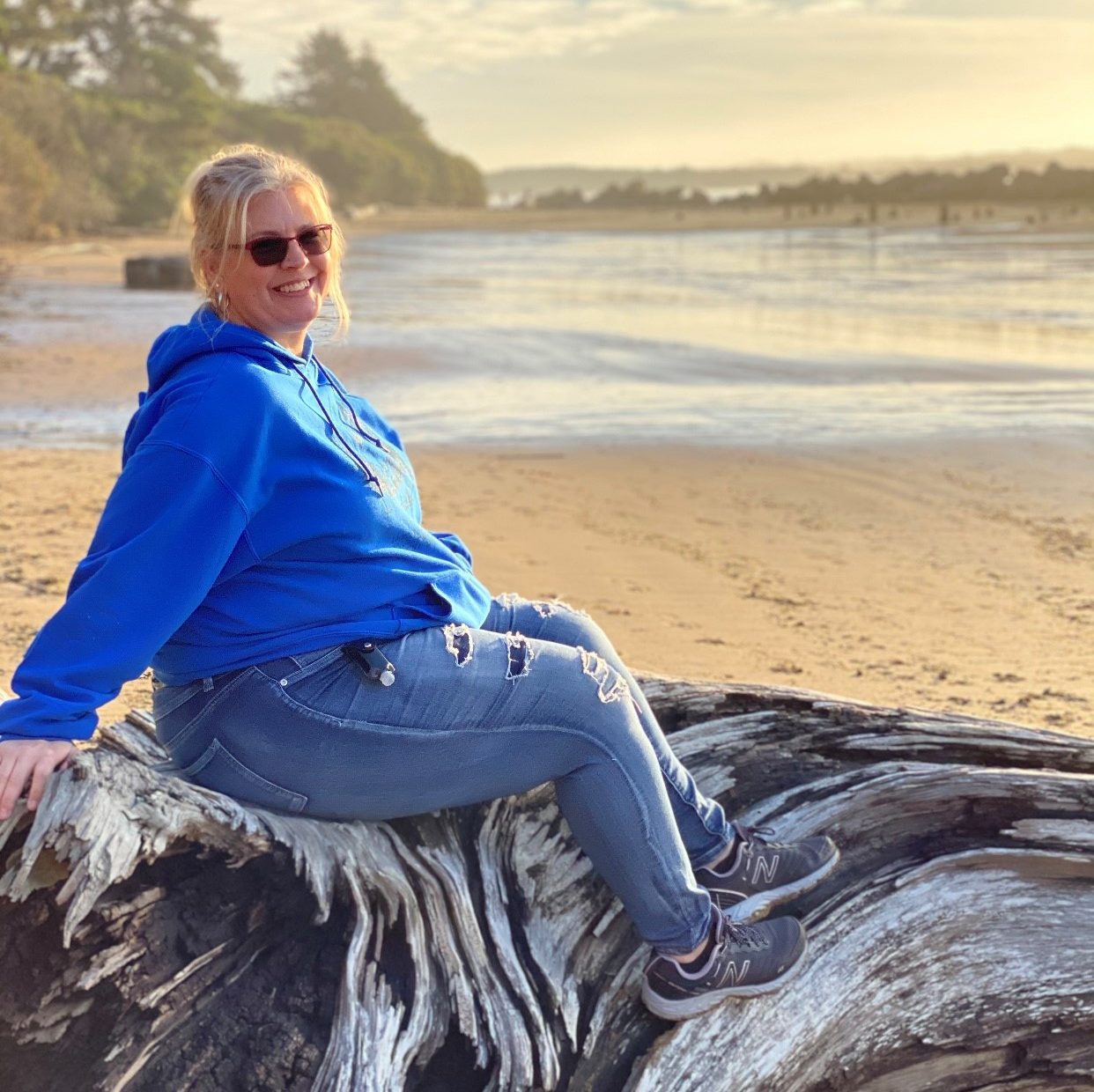 blonde woman sitting on fallen tree and taking photo with coast in the background