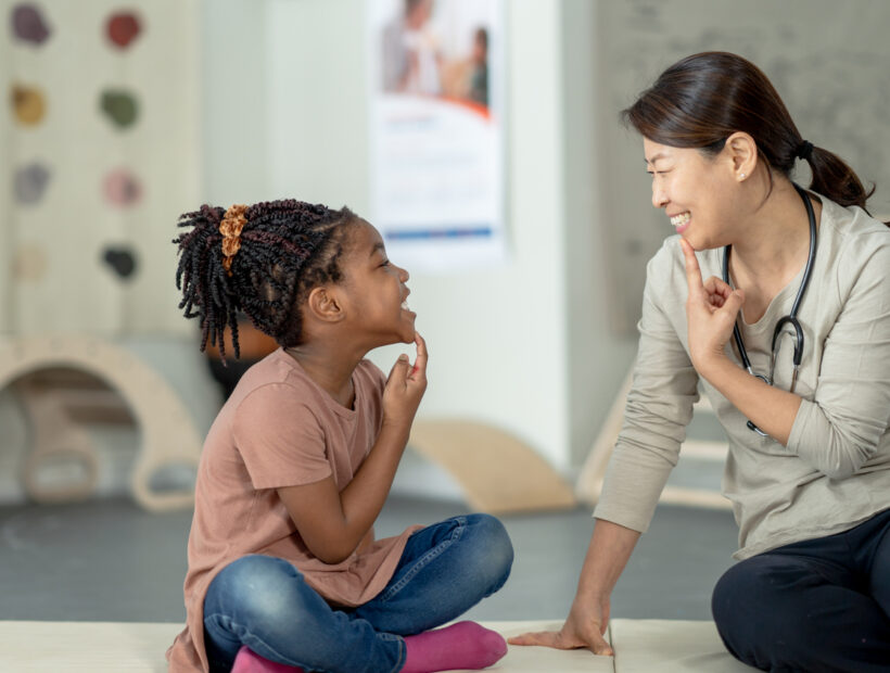 A female therapist sits beside her young patient on the floor as they work on some speech therapy exercises. They are each dressed casually and have their hands on their chins as they work through phonetical sounds. Small white letter tiles can be seen laying out in front of them from a previous exercise.