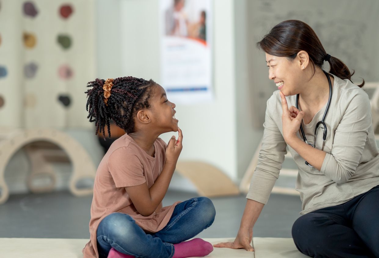 A female therapist sits beside her young patient on the floor as they work on some speech therapy exercises. They are each dressed casually and have their hands on their chins as they work through phonetical sounds. Small white letter tiles can be seen laying out in front of them from a previous exercise.